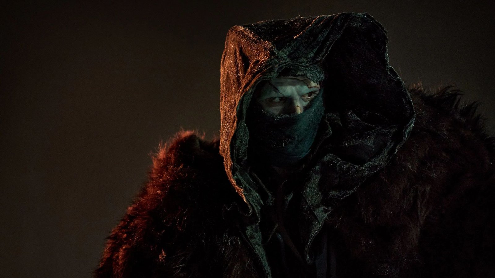 Um personagem encapuzado, com o rosto parcialmente coberto por um tecido escuro, olhando para o lado em um ambiente sombrio. Ele veste um manto pesado com pele escura e aparência desgastada.